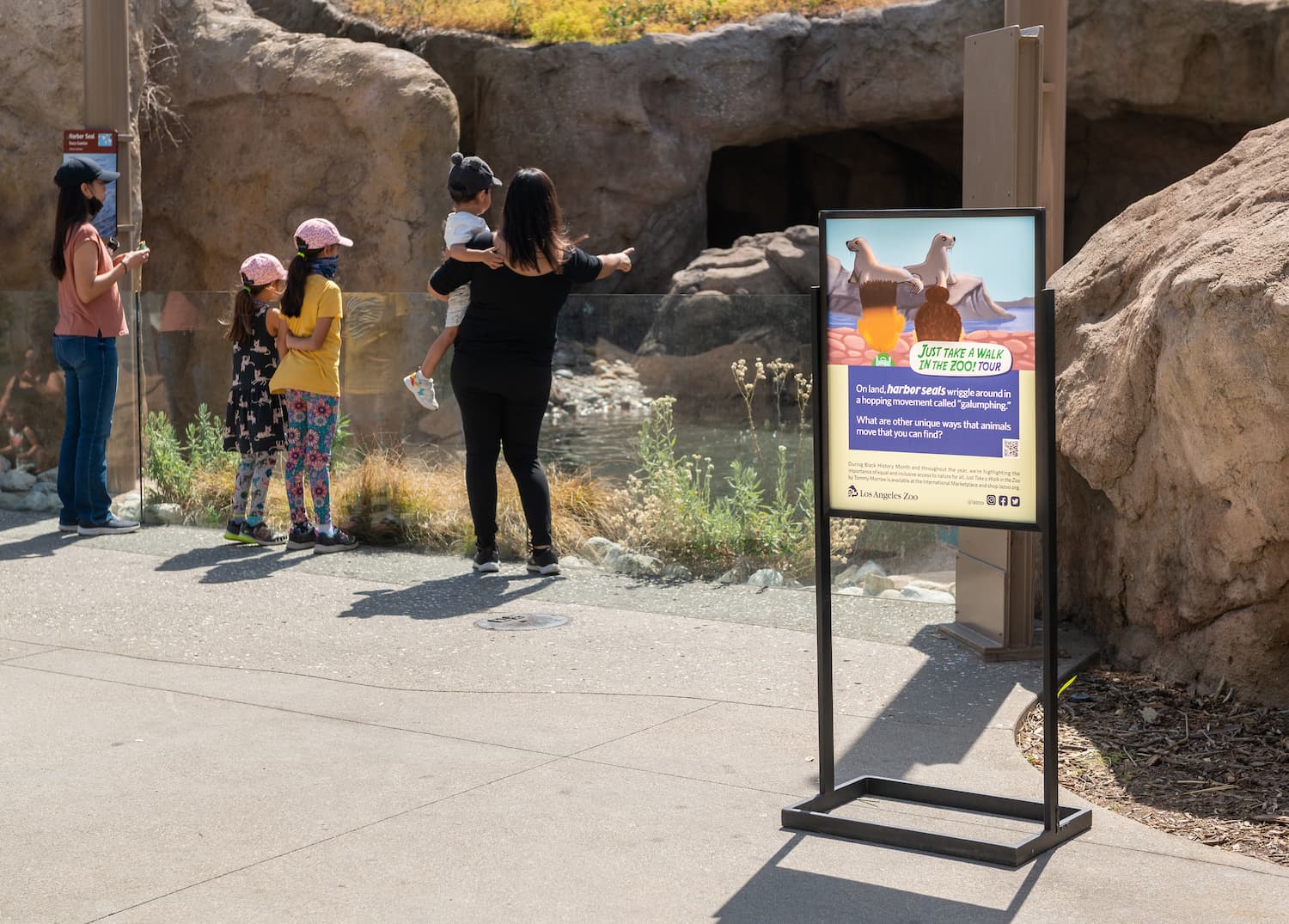 Guests take in views of the Sea Life Cliffs habitat, where a sign inspired by the children's book "Just Take a Walk in the Zoo" is displayed. The sign features an illustration from the book depicting two African American children exploring a zoo. The copy reads, "On land, harbor seals wriggle around in a hopping movement called 'galumphing.' What are other unique ways that animals move that you can find?"