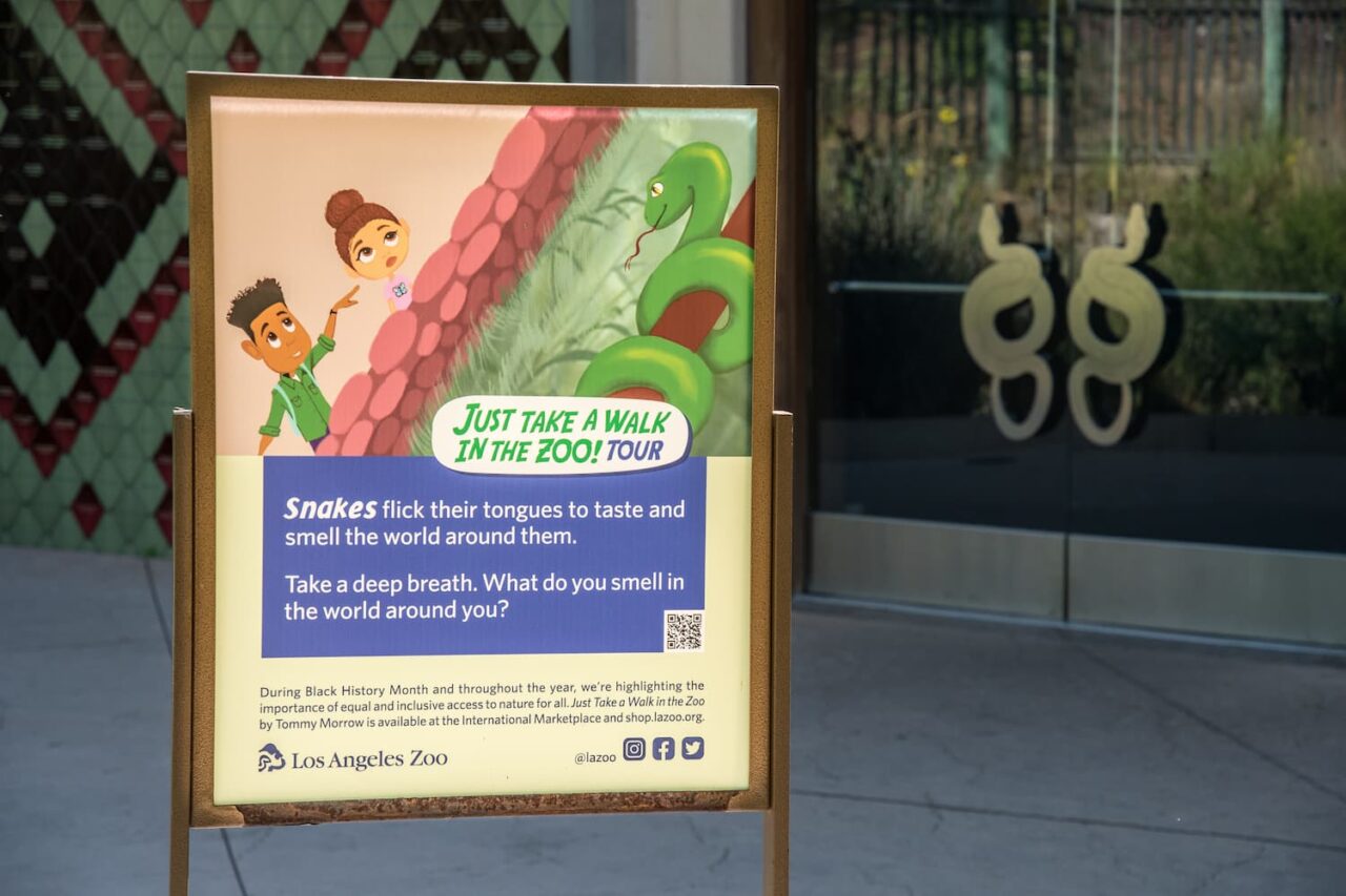 A sign inspired by the children's book "Just Take a Walk in the Zoo" is displayed in front of the LAIR. The sign features an illustration from the book featuring two African American children viewing a snake at a zoo. The copy reads, "Snakes flick their tongues to taste and smell the world around them. Take a deep breath. What do you smell in the world around you?"
