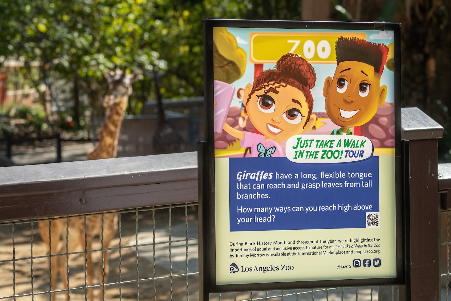 A sign inspired by the children's book "Just Take a Walk in the Zoo" is displayed in front of the Masai giraffe habitat. The sign features an illustration from the book featuring two African American children exploring a zoo. The copy reads, "Giraffes have a long, flexible tongue that can reach and grasp leaves from tall branches. How many ways can you reach high above your head?"