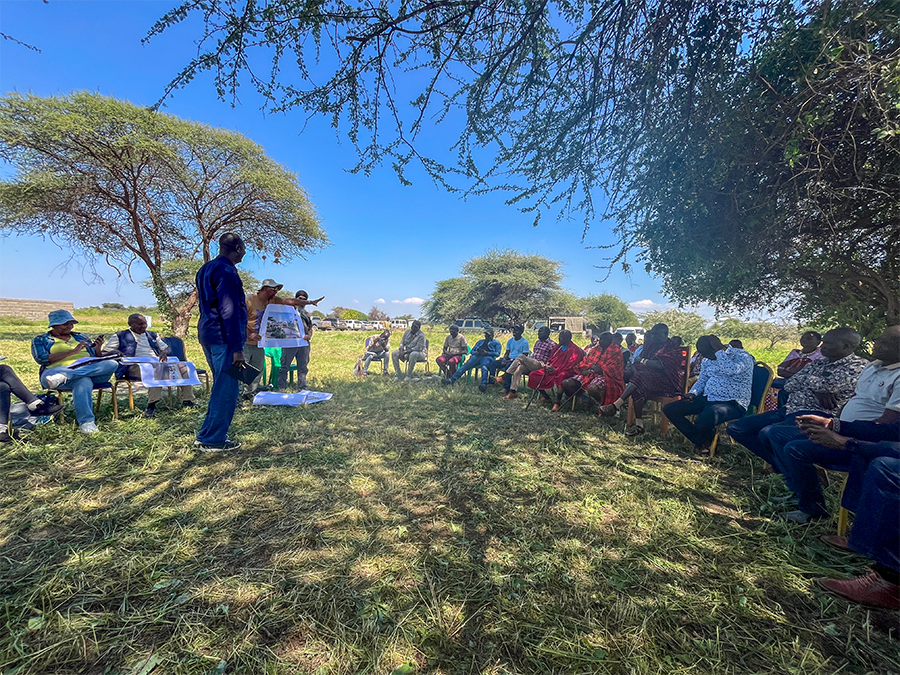 A large group sits in a shaded circle underneath trees, listening to a speaker who holds up a map.
