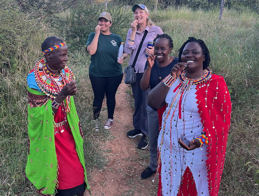 Five women in Kenya stand along a dirt path, smiling while demonstrating traditional teeth cleaning using small sticks.