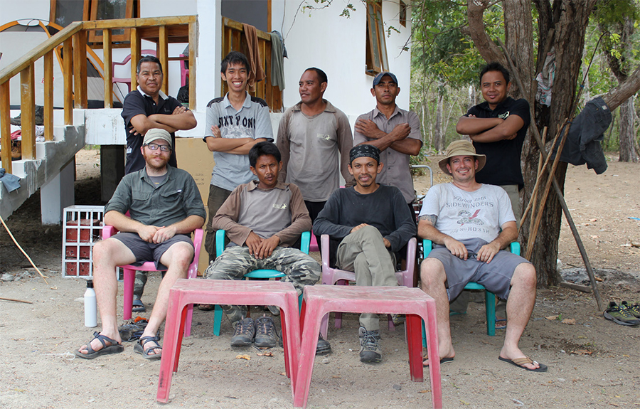 Nine middle-aged men pose for a group shot on colorful plastic chairs against a rustic background.