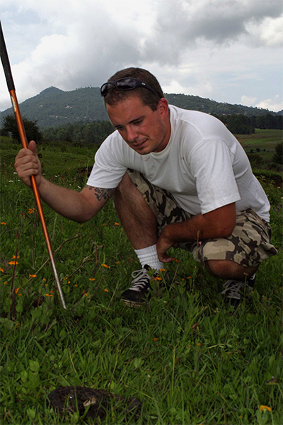 Ian Recchio crouches in a grassy field over a coiled-up rattlesnake.