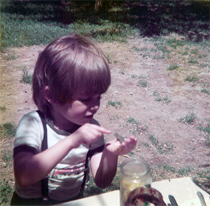 Ian Recchio, as a little boy with suspenders and a bowl haircut, holds a lizard in one hand and gently reaches to touch it with the other.