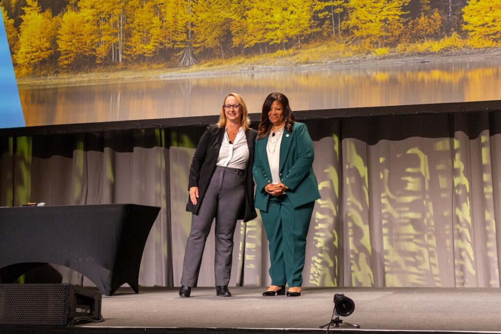 : Two business women in suits stand on a stage, posing for a photo.