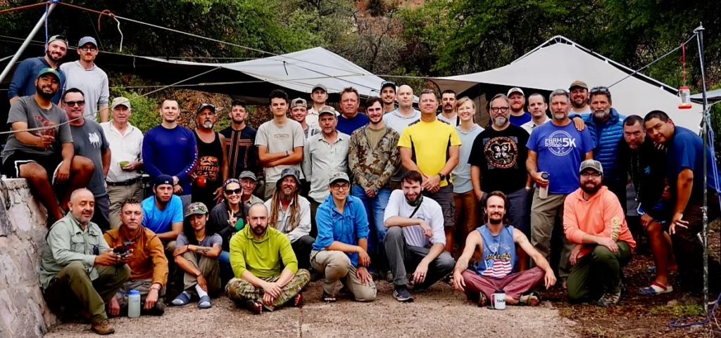 Group photo of individuals participating in "Project Obscurus" the new mexico ridge-nose rattlesnake conservation project. The group consists of diverse people, some wearing outdoor gear, standing and sitting in front of tents and natural vegetation. The atmosphere appears casual and collaborative, reflecting a shared interest in conservation or fieldwork.