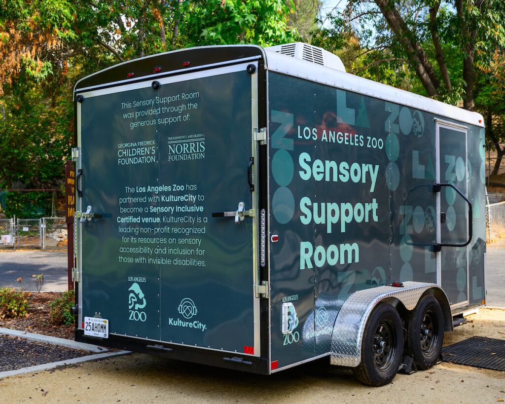 A green trailer with Zoo logos and designs sits parked in front of a grove of trees.