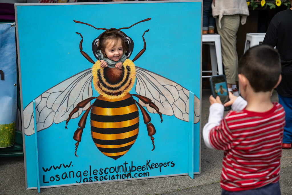 A child smiles through a cutout of a giant bee as another child takes a picture.