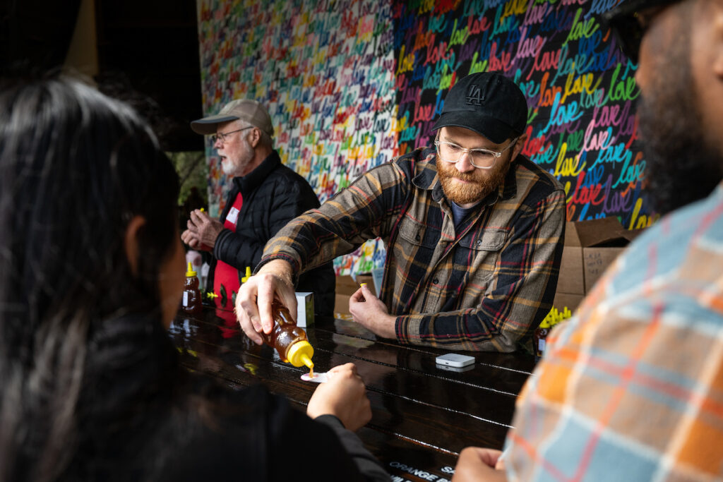 A vendor in a plaid shirt pours honey samples for visitors at a tasting station.