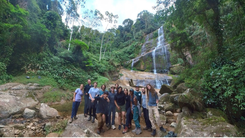 A group of students poses on a rocky outcrop with a forest waterfall in the background.