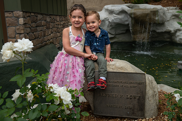 a young girl in a pink dress and a toddler boy in a blue shirt and green pants sit atop a rock with a plaque recognizing donor Alfred P Lowy