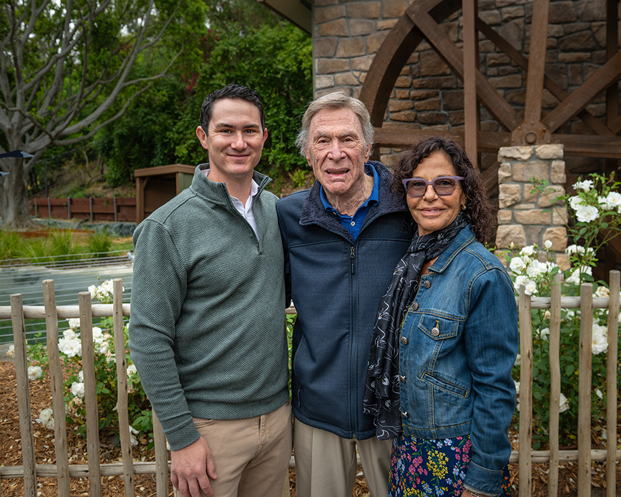 An older man poses with his arms around his adult son and daughter in front of a water wheel and pond with trees and white flowers in the background.