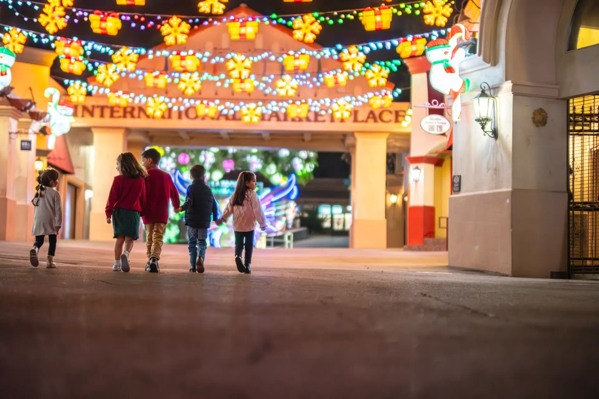 children holding hands and walking beneath strands of string lights